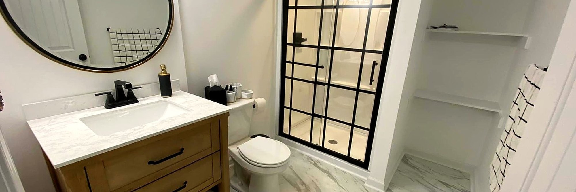 Finished bathroom with a stone-topped vanity, round mirror and a black-framed glass shower.